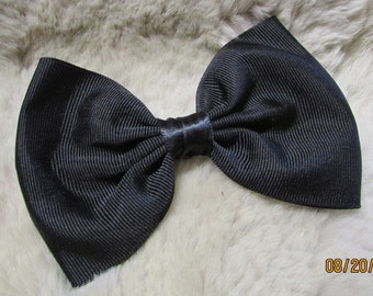 May include: A large, black bow made of ribbed fabric. The bow is tied in the center with a satin ribbon. The bow is a solid black color and is set against a light-colored, textured background.