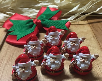 May include: A collection of small Santa Claus figurines with red suits, hats, and white beards. Each figurine is holding a brown sack. A red bow with green ribbon accents is in the background. The figurines are arranged on a wooden surface.
