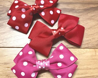 May include: Three ribbon hair bows in red and pink. The red bows have white polka dots, while the pink bow has white polka dots. The bows are arranged on a wooden surface.