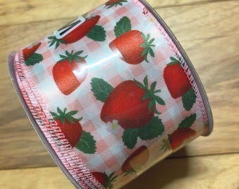 May include: A roll of decorative ribbon featuring a repeating pattern of red strawberries with green leaves on a pink and white gingham background. The ribbon has a pink stitched edge and is ideal for crafts and gift wrapping.