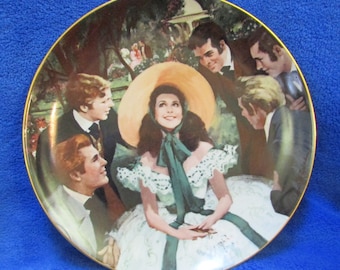 May include: A decorative ceramic plate featuring a painting of a woman in a white dress and a large straw hat surrounded by four men. The plate has a gold rim and a green background.