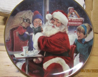 May include: A vintage Christmas-themed collectible plate featuring Santa Claus in a red suit with a white beard, a young boy in a blue and white outfit, and a young girl in a blue and white outfit. The plate has a gold rim and a blue background with white snowflakes. The text "15¢ McDonald's Hamburgers" is visible in the background.