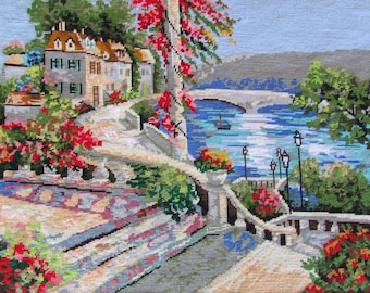 May include: A cross-stitch embroidery depicting a picturesque scene of a stone bridge over a blue river, with colorful flowers and a small village in the background.