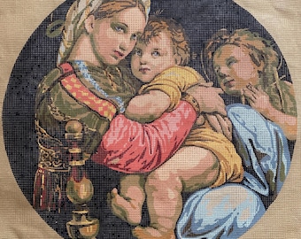 May include: Needlepoint artwork depicting a Renaissance-style scene. A woman in a headscarf cradles a child, with another figure to the side. The artwork features a dark blue background and a circular shape.