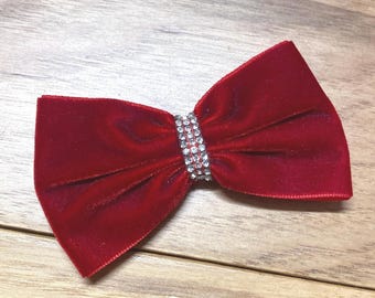 May include: A deep red velvet bow with a sparkling rhinestone center. The bow is tied in a classic shape, with a soft, textured appearance. The rhinestone band adds a touch of glamour to this hair accessory.
