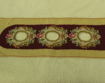 May include: A beige canvas with a burgundy border featuring three embroidered floral designs with circular cutouts. The design includes pink, green, and white flowers.