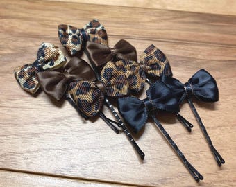 May include: A collection of hair bows in various styles and colors. The bows include leopard print, brown satin, and black satin bows, all attached to bobby pins. The bows are arranged on a wooden surface.