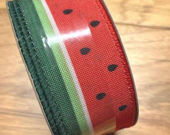 May include: A roll of decorative ribbon featuring a watermelon design. The ribbon has a red background with black seed details, green and white stripes, and a green edge. The ribbon is on a wooden surface.