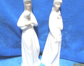 May include: Two white porcelain figurines of women, each holding a small animal. The woman on the left is holding a small dog, and the woman on the right is holding a small bird. Both figurines are wearing long white dresses.