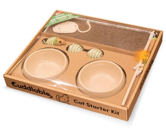May include: A cardboard box with a clear plastic window showing a cat starter kit. The kit includes two beige ceramic bowls, a scratching post, a toy mouse, and two green and white striped balls. The box is labelled "Cuddlable Cat Starter Kit".