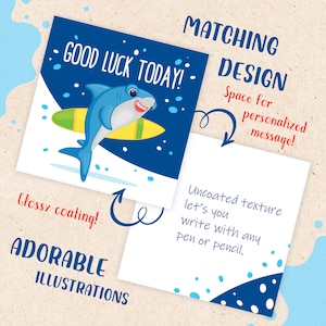May include: A blue and white card with a cartoon shark surfing on a yellow surfboard. The card has the text "GOOD LUCK TODAY!" and is designed for writing a personalized message. The card has a glossy coating and is paired with a blank card with a white background and blue and white polka dots.