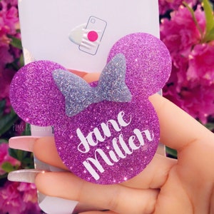 Pode incluir: Um suporte de telefone PopSockets roxo brilhante em forma de orelhas da Minnie Mouse com um laço de glitter prateado. O nome "Jane Miller" está escrito em branco. O PopSockets está em um fundo branco com o logotipo PopSockets.