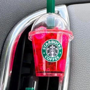 May include: A Starbucks-branded car air freshener shaped like a pink drink. The clear plastic cup has a green straw and a Starbucks logo. The drink appears to have fruit pieces inside.