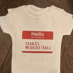 以下が含まれることがあります： 赤色の名前タグに「Hello my name is Cranjis Mcbasketball」と書かれた白いベビーロンパースです。