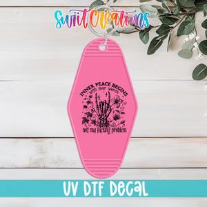 May include: Pink motel-style keychain with the text "INNER PEACE BEGINS with four words: not my f*cking problem" and a black skeleton hand graphic. The keychain has a silver ring and the words "UV DTF DECAL" at the bottom.