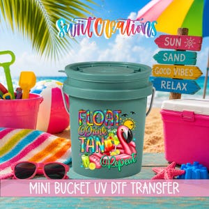 May include: A sage green plastic bucket with a lid, featuring a colorful graphic print that reads "FLOAT Drink TAN Repeat" with a pink flamingo float, tropical drinks, and a sun. The bucket is surrounded by beach toys, sunglasses, and a towel.