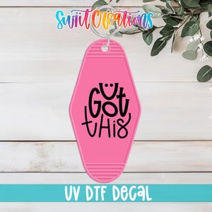 Puede incluir: Llavero rosa estilo motel con la frase "Got This" en escritura negra. El llavero tiene un anillo de metal en la parte superior y las palabras "UV DTF DECAL" en blanco. El fondo es una superficie de madera blanca.