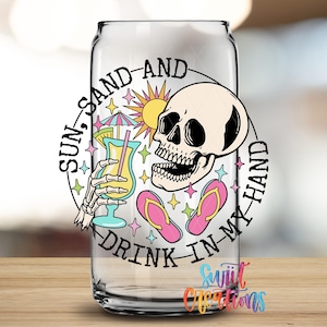 A glass with a colorful design featuring a skull, sunglasses, a beach umbrella, and a cocktail glass, along with the text "Sun, sand, and drink in my hand".