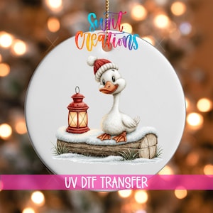 Puede incluir: Un adorno redondo de cerámica blanca con una oca de dibujos animados con un gorro de punto rojo y blanco, sentada sobre un tronco cubierto de nieve junto a una linterna roja brillante. El texto Swift Creations y UV DTF TRANSFER es visible.