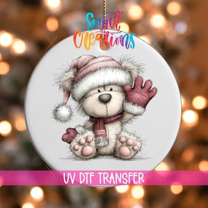 May include: A round white ceramic ornament featuring a cartoon bear wearing a pink winter hat and scarf with matching mittens. The bear is waving one hand. The text "Swift Creations" appears at the top, and "UV DTF TRANSFER" is at the bottom.