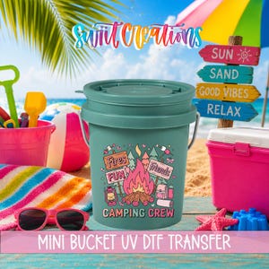 May include: A teal mini bucket with a camping-themed graphic, including a campfire, trees, and the words "Fires, Fun, Friends, Camping Crew." The bucket is on a sandy beach with beach accessories and the text "MINI BUCKET UV DTF TRANSFER."