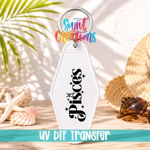 May include: A white, diamond-shaped motel-style keychain featuring the black text "Pisces" oriented vertically, accented with small star symbols and a Pisces zodiac icon. The keychain includes a silver metal split ring at the top for attachment.