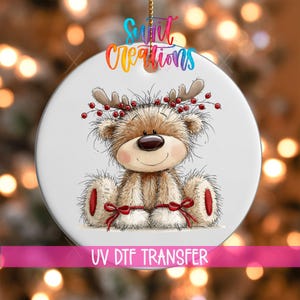 May include: A round white ceramic ornament featuring a fluffy brown teddy bear with reindeer antlers adorned with red berries. The bear has red bows on its feet and the text "Swift Creations" is printed at the top, with "UV DTF TRANSFER" below.