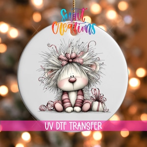 Könnte beinhalten: Ein rundes weißes Keramikornament mit einer bunten Illustration eines flauschigen Wesens mit rosa Schleife, das neben einem Geschenk und Wollknäueln sitzt. Der Text "Swift Creations" steht oben und "UV DTF TRANSFER" unten auf dem Ornament.