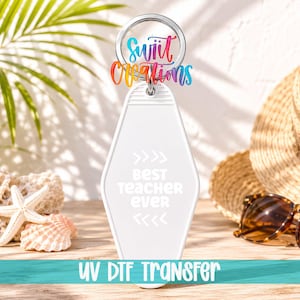 May include: A white, diamond-shaped motel-style keyring featuring a silver metal ring. The keyring is printed with the text 'BEST TEACHER EVER' in white, centred between two sets of chevron arrows. Above the ring, a colourful logo reads 'swiit creations'.