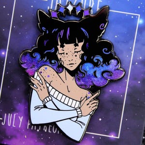 May include: Enamel pin depicting a celestial woman with black cat ears, a star crown, and galaxy-themed hair and accents. The woman is wearing a light blue sweater. The pin is set against a purple background with the text "JULY".