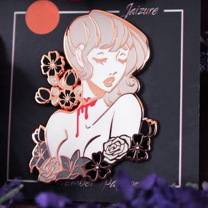 May include: Rose gold enamel pin featuring a woman with white hair and a red drip down her neck. The woman is surrounded by black and white flowers. The pin says "Jaizure" at the top and "Remember" at the bottom.