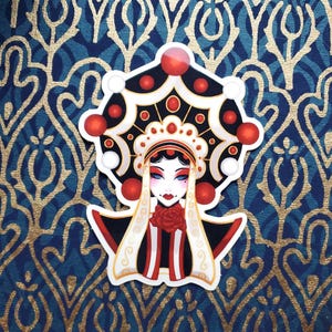 May include: A sticker featuring a stylized illustration of a woman wearing a traditional Chinese opera costume. The costume is black, red, and white with intricate details. The woman has black hair and red lips.