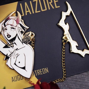 Puede incluir: Un pin de metal dorado y un marcapáginas a juego. El pin presenta una ilustración en blanco y negro de una mujer con cuernos. El marcapáginas tiene un diseño de arco y flecha con esmalte blanco. Ambos están conectados por una cadena dorada.
