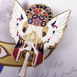 Puede incluir: Pin de esmalte dorado con un ángel blanco con alas, sosteniendo una espada. El ángel tiene una cara detallada con una corona de ojos y una estrella. El fondo es una tela blanca con un diseño de ojo dorado y naranja.