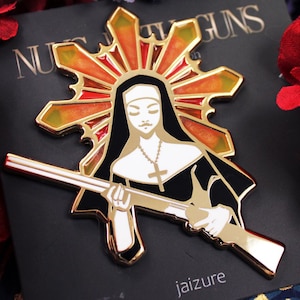May include: Gold-toned enamel pin featuring a nun holding a rifle, with a halo of stained glass behind her. The text "Nuns With Guns" is visible in the background.