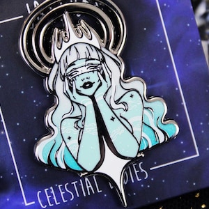 May include: A silver-toned enamel pin featuring a celestial figure with flowing white hair and a crown. The figure has a light blue skin tone and is holding their face. The pin is set against a dark blue background with the text "CELESTIAL NOTES".