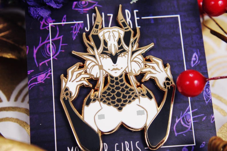 May include: A gold-toned metal enamel pin featuring a stylized character with white hair, horns, and pointed ears. The character wears a black scale-patterned collar and black gloves, with hands raised near the head, set against a dark purple backing card.