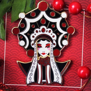 May include: A black and white enamel pin featuring a stylized woman with a red and gold headdress. The pin is set against a red background with a wave pattern.