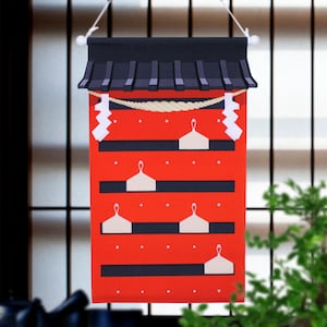 May include: A red and black paper shrine with a rope and white paper charms hanging from the top. The shrine has five shelves with white paper charms on each shelf.