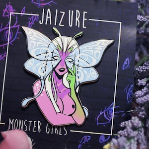 以下が含まれることがあります： 蝶の羽根とマスクをつけた女性を描いた青と白のエナメルピン。ピンは「JAIZURE」と「MONSTER GIRLS」の文字が書かれた黒い背景の上にあります。