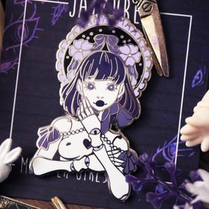 May include: Purple and white enamel pin featuring a girl with a crown of flowers and a skull in her hand. The pin has a detailed design and a glossy finish.