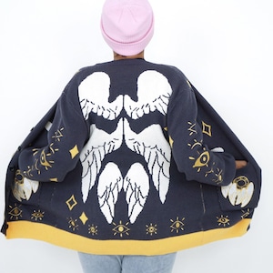 May include: Navy blue cardigan with a white angel wing pattern on the back. The sweater features gold star and eye details, and a yellow trim. The person in the image is wearing a pink beanie.