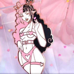 May include: A pink and black enamel pin featuring a stylized illustration of a woman in lingerie. The pin has a rose gold outline and includes heart-shaped details. The woman has pink and black hair and is holding up a heart.