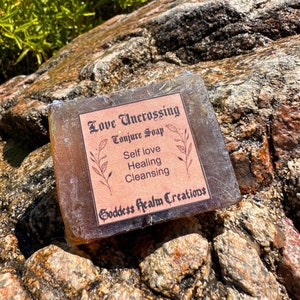 May include: A square bar of soap with a brown hue, resting on a textured rock surface. The soap has a label with the words "Love Uncrossing Conjure Soap", "Self love, Healing, Cleansing", and "Goddess Realm Creations". Green foliage is in the background.