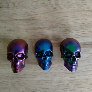 Könnte beinhalten: Drei schädelartige, schillernde Objekte mit metallischem Finish. Die Schädel zeigen eine Reihe von Farben, darunter Lila, Blau und Grün, und sind auf einer Holzoberfläche angeordnet. Die Schädel sind ein dekorativer Artikel.