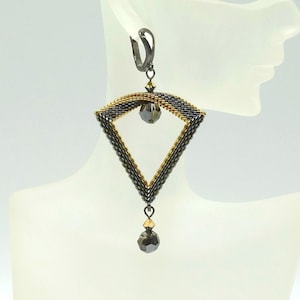 Könnte beinhalten: Ein einzelner, geometrischer Ohrring in rautenförmiger Form, gefertigt aus kleinen, dunklen Perlen. Der Ohrring hat einen goldfarbenen oberen Teil und eine herabhängende, facettierte Perle. Das Design wird auf einem Schaufensterkopf präsentiert.