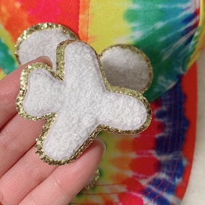 以下が含まれることがあります： 飛行機の形をした、金色のグリッターの縁取りが付いた白いふわふわのパッチ。パッチはタイダイ柄の布製の帽子に取り付けられています。