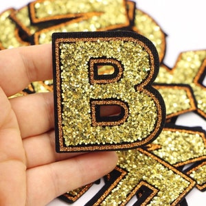 May include: Gold glitter letter B patch with a black outline and orange trim. The patch is made of fabric and has a sparkly finish.