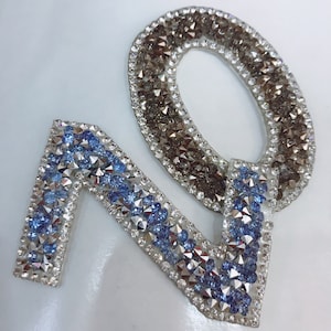 Pode incluir: Um par de patches para passar a ferro em forma dos números "7" e "0". O número "7" é feito de strass azul e prata. O número "0" é feito de strass prata e preto.