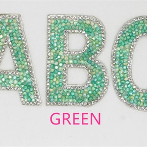 Könnte beinhalten: Drei grüne Strass-Buchstaben, A, B und C, mit silbernem Rand. Die Buchstaben befinden sich auf weißem Hintergrund mit dem Wort "GRÜN" darunter.